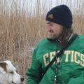 A slightly scruffy person with a bent nose and long hair, wearing a dark blue winter cap and a green Celtics jacket, looks to his left toward a good dog.