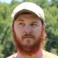 A man with a full reddish beard and beige cap looks to the side outdoors with a blurred green background.
