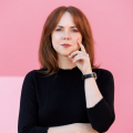 Woman in a black mock turtleneck stands in front of a pink background: she has red hair and pink lipstick