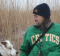 A slightly scruffy person with a bent nose and long hair, wearing a dark blue winter cap and a green Celtics jacket, looks to his left toward a good dog.