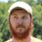 A man with a full reddish beard and beige cap looks to the side outdoors with a blurred green background.