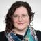 Photo of a nonbinary person with pale skin, dark curly hair that is long on one side and short on the other. They are wearing gold-rimmed glasses and a colorful scarf. They are smiling slightly at the camera. 