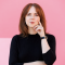Woman in a black mock turtleneck stands in front of a pink background: she has red hair and pink lipstick