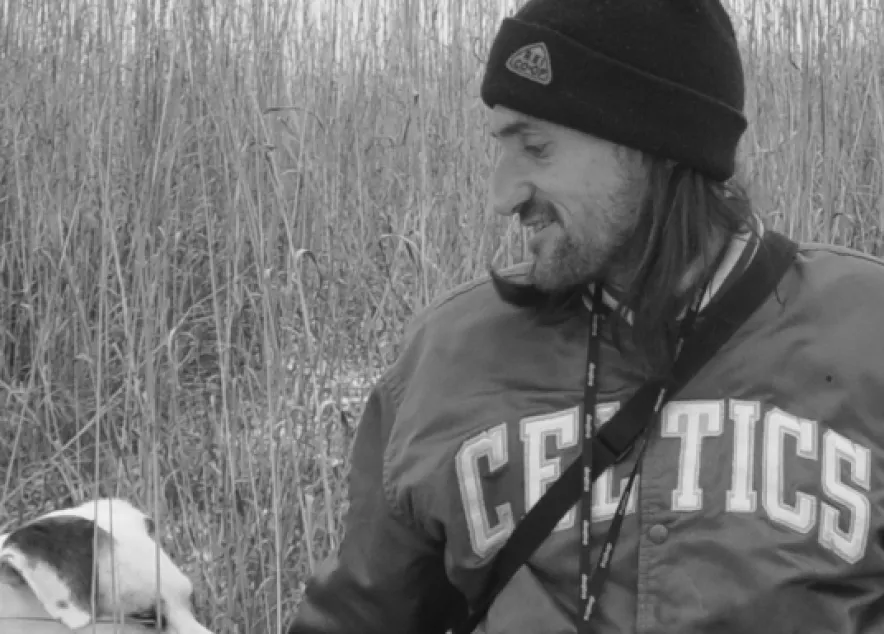 A slightly scruffy person with a bent nose and long hair, wearing a dark blue winter cap and a green Celtics jacket, looks to his left toward a good dog.