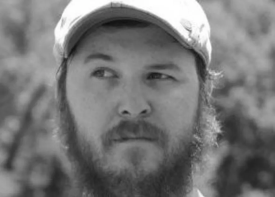 A man with a full reddish beard and beige cap looks to the side outdoors with a blurred green background.