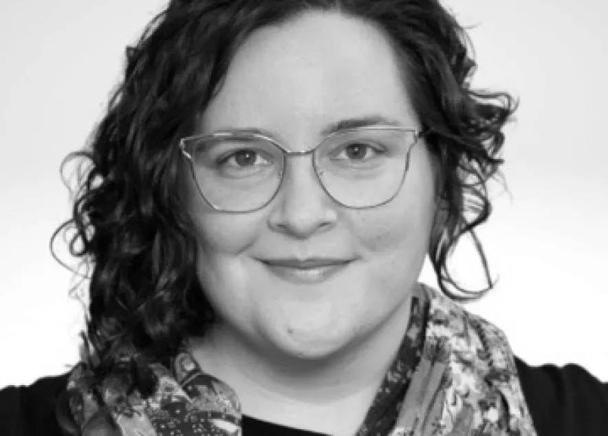 Photo of a nonbinary person with pale skin, dark curly hair that is long on one side and short on the other. They are wearing gold-rimmed glasses and a colorful scarf. They are smiling slightly at the camera. 