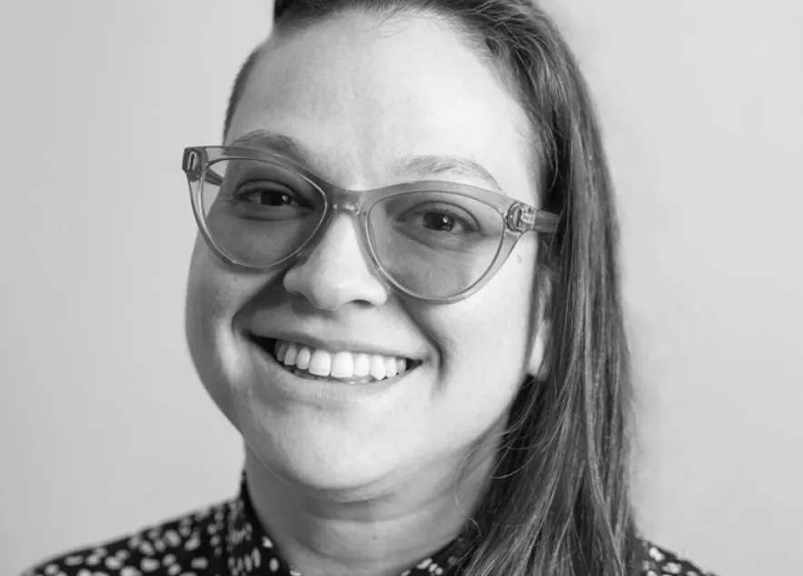 White woman in her late thirties wearing purple glasses, jewelry, a button-down dress, and a smile.