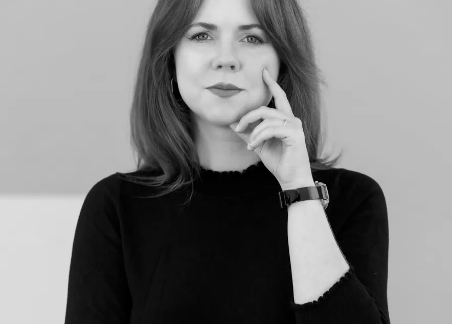 Woman in a black mock turtleneck stands in front of a pink background: she has red hair and pink lipstick
