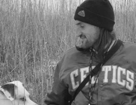 A slightly scruffy person with a bent nose and long hair, wearing a dark blue winter cap and a green Celtics jacket, looks to his left toward a good dog.