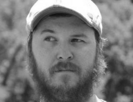 A man with a full reddish beard and beige cap looks to the side outdoors with a blurred green background.