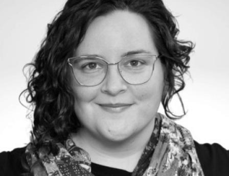 Photo of a nonbinary person with pale skin, dark curly hair that is long on one side and short on the other. They are wearing gold-rimmed glasses and a colorful scarf. They are smiling slightly at the camera. 