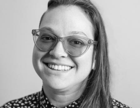 White woman in her late thirties wearing purple glasses, jewelry, a button-down dress, and a smile.